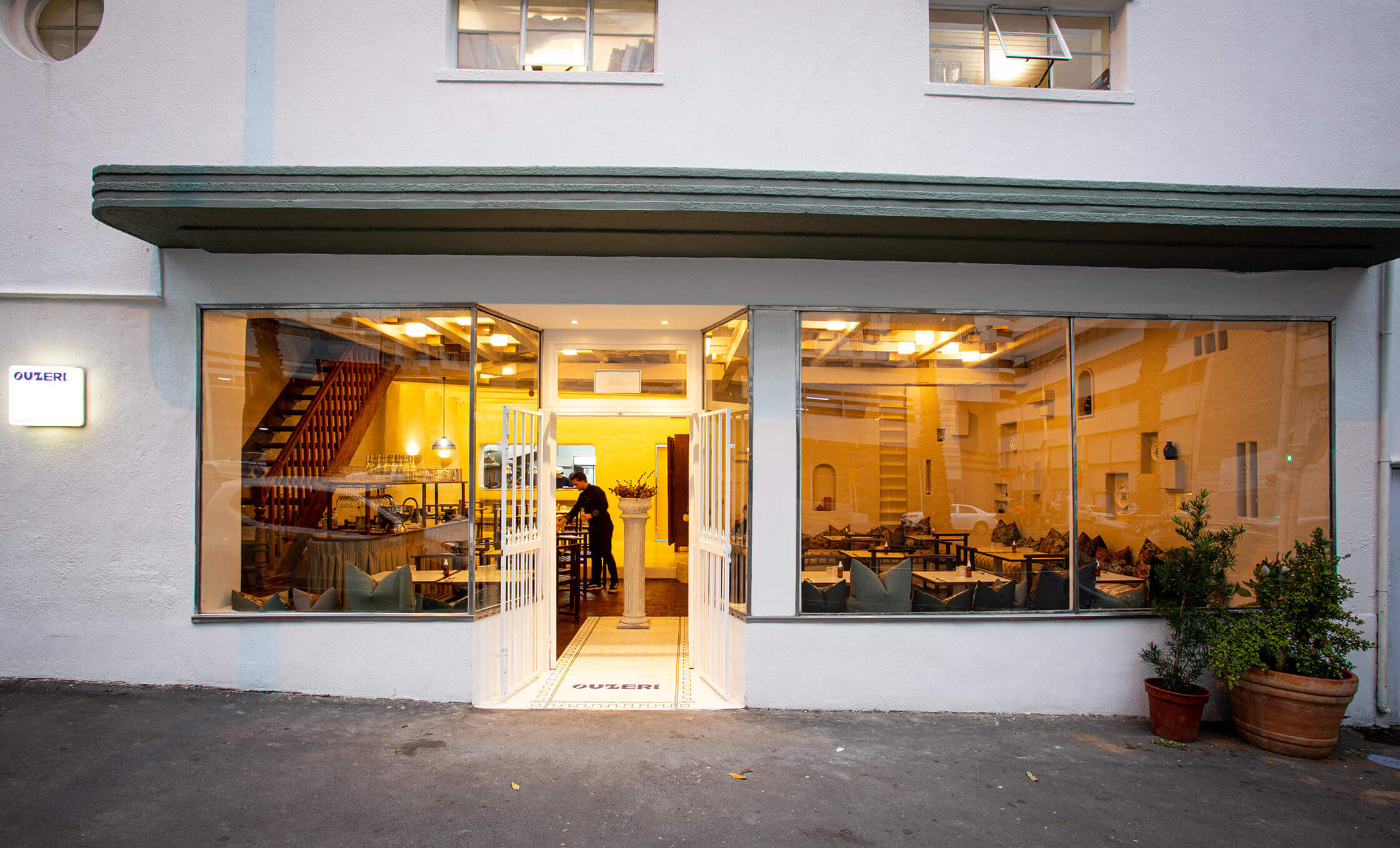 Ouzeri restaurant warmly lit with large glass windows and open white double doors.